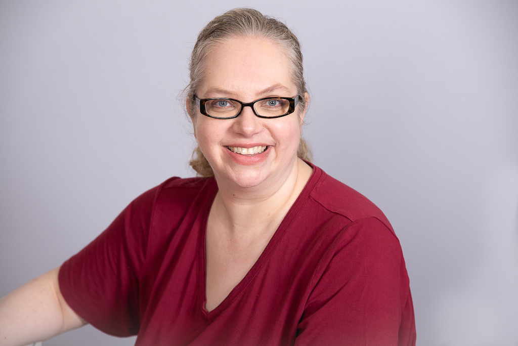 This is a professional headshot of Ann Dreolini. Ann is wearing a red shirt, black glasses, and is looking straight at the camera. Behind her is a plain grey backdrop.