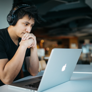 photo of Asian man in a black shirt with over the ear black headphones. He is sitting at a table looking intently at a MacBook. the background is blurred and can be assumed he is in an empty coffee shop.