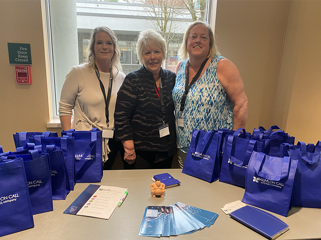 Representatives of H.I.M. on Call, Jodi McMullin, Debra Tetreault, and Tammy Hutt, huddled together for a photograph in front of a table with blue souvenir bags.